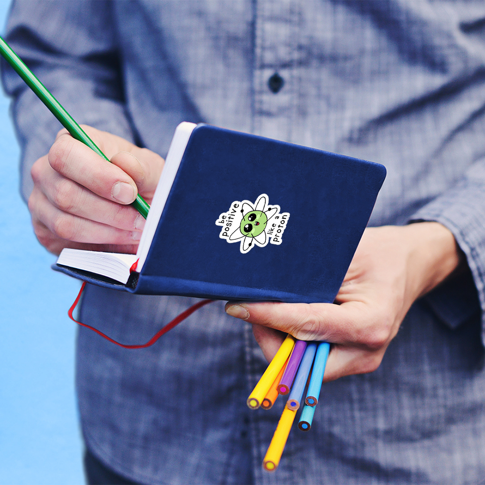 Cute science stickers of a smiling atom on a blue notebook held by person writing with green pencil