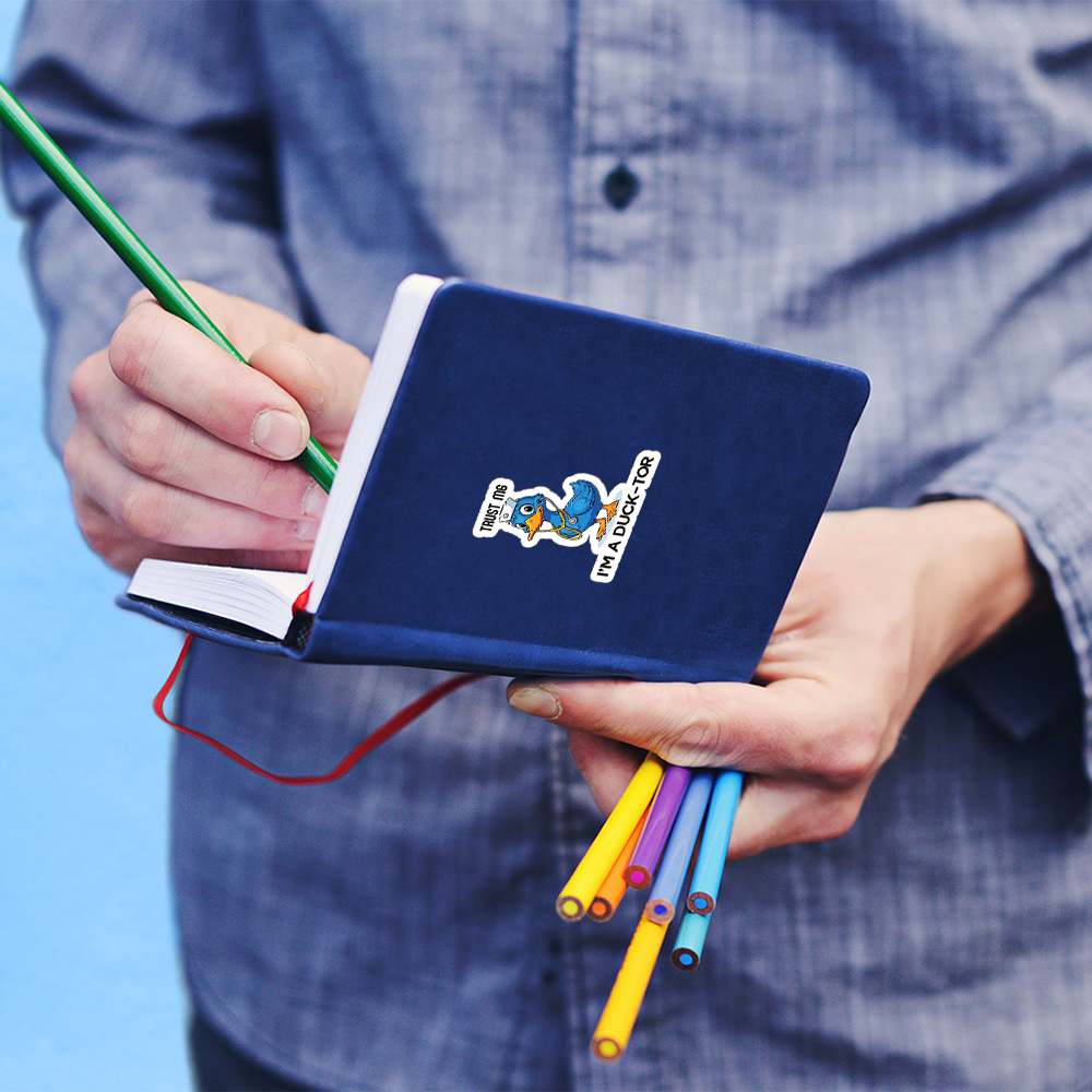 Funny duck doctor sticker on a blue notebook held by a person writing with a green pencil